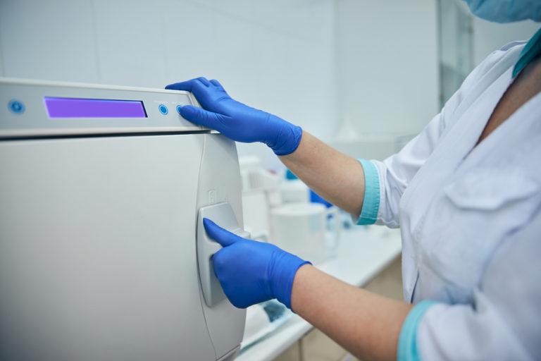 Close up image of woman hands in blue gloves while touching sensor panel of big sterilizer