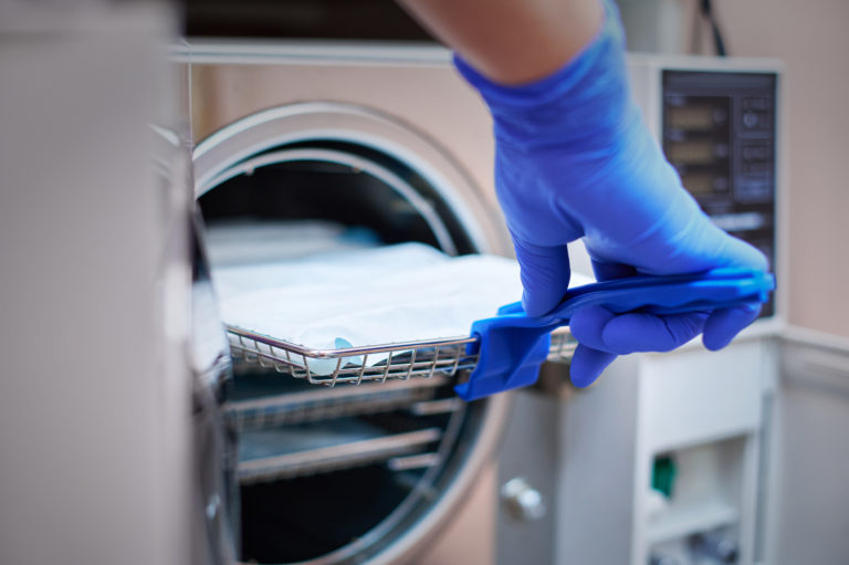 Dentist in blue latex gloves is taking a sealed dental stainless steel instrument from an autoclave sterilizer.